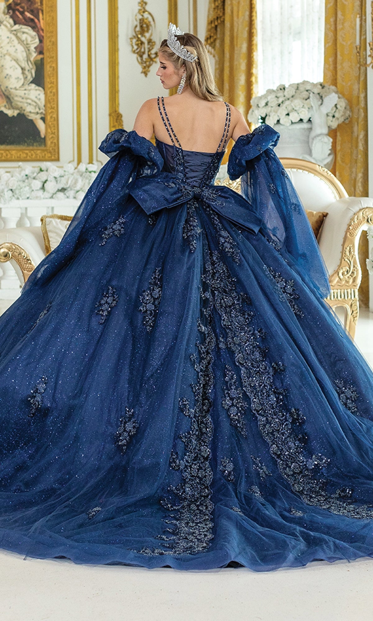 A woman in the Blue Quinceanera Dress 5037 by Dancing Queen, featuring blue glitter, floral embellishments, and a large bow, stands in an ornate gold-accented room with her back to the camera.